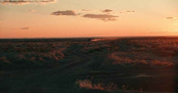 Distant cinematic shot of riders with a donkey cart at sunset, their silhouette framed by orange backlit dust rising from the desert road in Namibia.