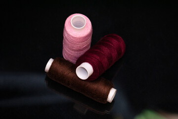 Three sewing thread spools in burgundy, pink and brown colors on a black background. Concept of tailoring, handmade craft, textile materials and fashion design accessories.