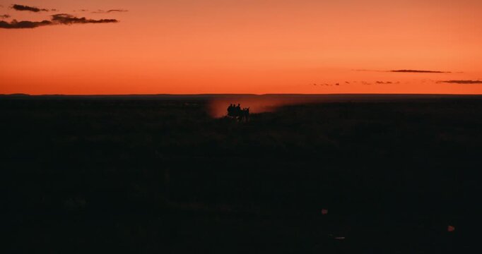 Distant cinematic shot of riders with a donkey cart at sunset, their silhouette framed by orange backlit dust rising from the desert road in Namibia.