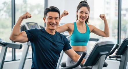 Fototapeta premium Smiling Asian man and woman flexing biceps on exercise machines during workout in a bright gym