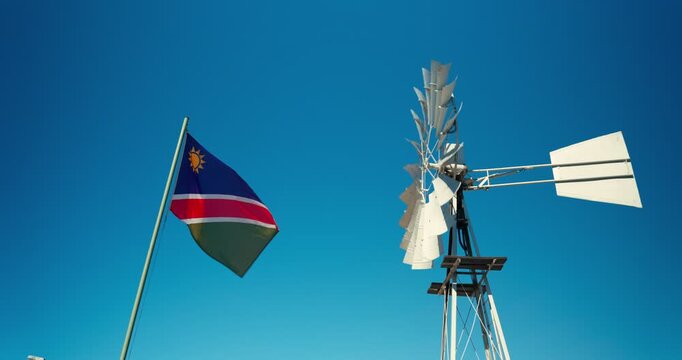Namibian flag flutters in the wind beside a spinning windwheel, symbolizing renewable energy and rural life, set against a clear blue sky in the Namibian desert.