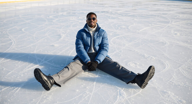 Happy African American man sitting on ice rink. Stylish winter fashion with blue puffer jacket and sunglasses. Copy space