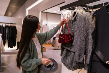 Young brunette woman shopping for Christmas season accessories, comparing glitter bags and matching them with a festive tweed outfit in a fashion store