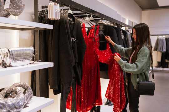 Young brunette woman choosing a red sequin dress during festive Christmas shopping, exploring holiday outfits and sparkling party looks in a fashion store