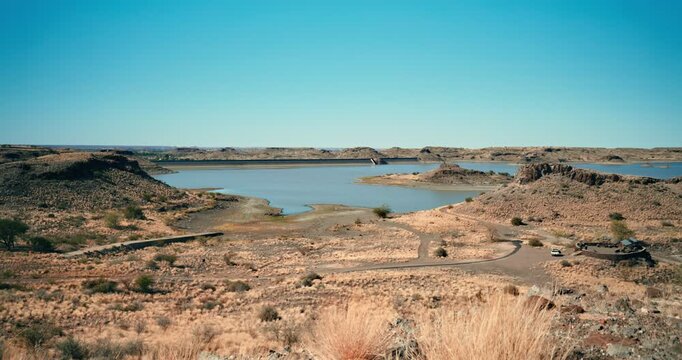 Tall dry grass moves in the desert wind, with a distant water reservoir in the background, creating a stark, dystopian landscape under soft daylight in Namibia.