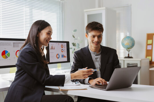 Asian business professionals discussing financial data on laptop and computer monitors, working together in a modern office setup - Powered by Adobe