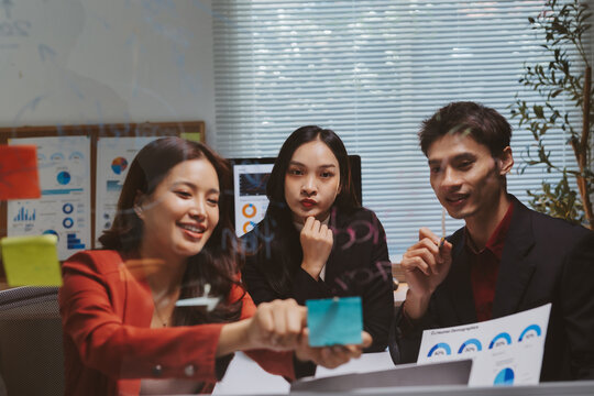 Young Asian business professionals brainstorming ideas and analyzing charts on a glass board during a corporate meeting