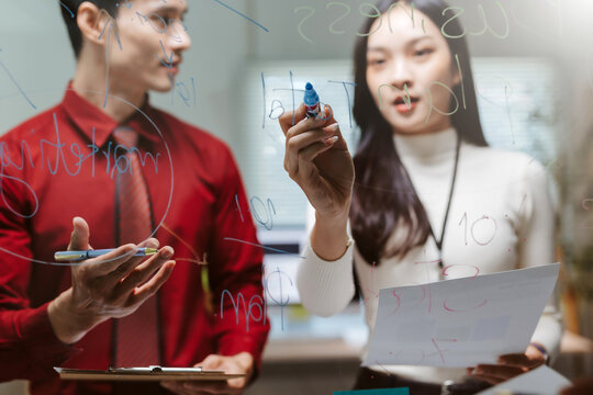 Asian business people collaborating and brainstorming, writing ideas and strategies on a glass whiteboard during an office meeting