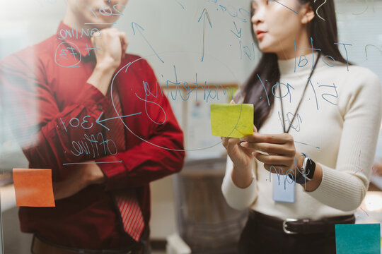 Business people planning and collaborating, writing on transparent glass board with colorful sticky notes during strategy meeting