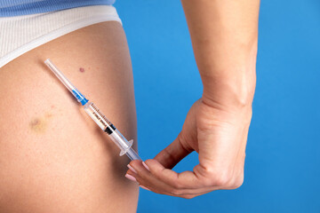 Close-up of a person administering a syringe injection into the thigh against a blue background,...