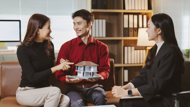 Asian couple smiling, reviewing house model with female real estate agent in an office, planning home ownership - Powered by Adobe
