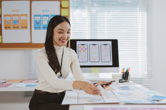 Asian woman smiles, creating mobile application wireframes, sketching user experience designs, and holding a smartphone in a modern office