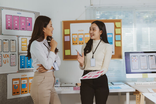 Two young asian women discussing and brainstorming user experience and interface design for a new mobile application in an office setting - Powered by Adobe
