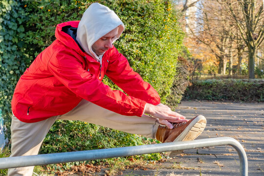 young man in red jacket stretching leg on park railing, warmup before run, solo training in cold air, discipline and small daily victories, public equipment for fitness routine, flexibility outdoors