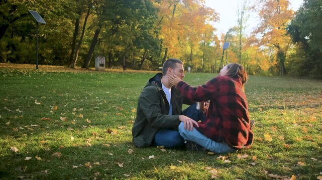 Romantic young multiracial couple enjoying a date in an autumn park, sitting on grass drinking tea.