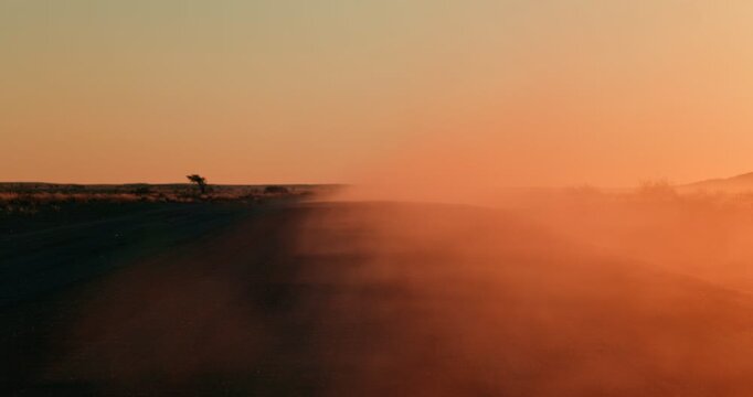 Orange dust rises over a gravel road cutting through Namibia&rsquo;s red desert at sunset, with warm glowing light reflecting beautifully in the drifting airborne dust.