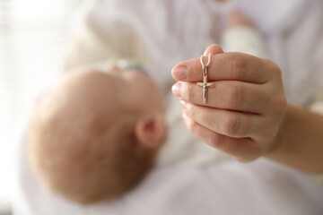 Sacrament of baptism. Woman holding little baby and gold cross indoors, selective focus