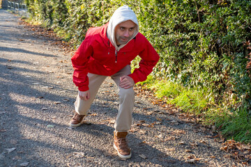 young man in red jacket legs and hamstrings, crouching on park path, performing pre run dynamic stretching, solo training in cold air, discipline and small daily victories, surrounded by green bushes