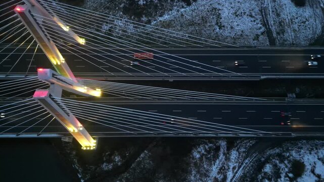 Aerial view captures cars driving along highway through the illuminated bridge at evening. Autos riding at speed road at cold season. Cable-stayed bridge spanning a river and snow-covered terrain