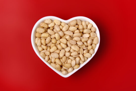 peeled pine nuts in a white heart-shaped bowl on a red background. top view.