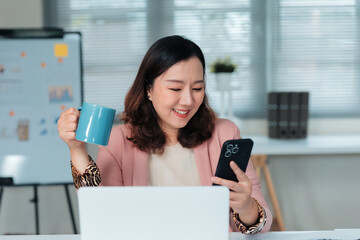 Asian businesswoman, smiling and happy, enjoying a coffee break while checking her smartphone in a professional office setting