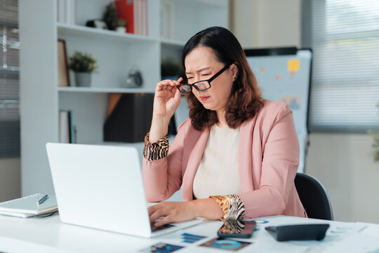 Mature Asian businesswoman needing reading glasses, struggling with vision and eyestrain while working on laptop in an office