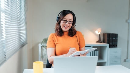 Smiling Asian woman wearing headset and glasses, taking notes during a video conference, webinar, or remote work meeting
