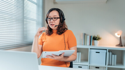 Asian woman wearing headset talking during virtual lesson or online meeting from home office, holding a digital tablet