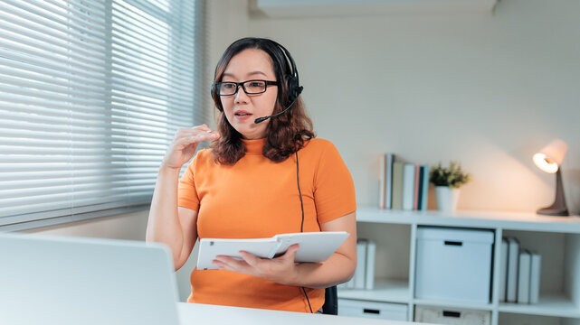 Asian woman wearing headset talking during virtual lesson or online meeting from home office, holding a digital tablet