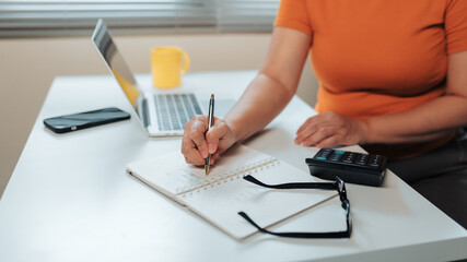 Woman managing personal finances and household budget, writing notes in a notebook at a desk with a laptop and calculator