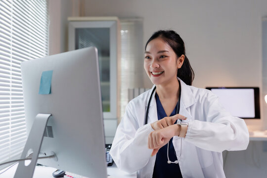 Young female doctor wearing a lab coat and stethoscope, looking at a computer screen and tapping smartwatch, providing online healthcare