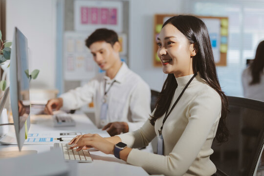 Professional Asian woman smiling while working on a computer in a modern office, collaborating with colleagues