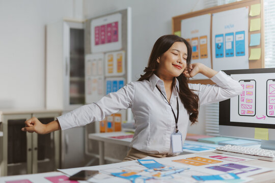 Asian woman stretching arms and relaxing at her modern office desk during a brief break from designing mobile apps