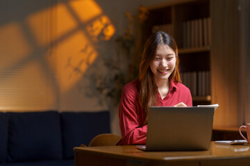 Young Asian woman smiling, doing remote work at home in the evening, using a laptop for online education
