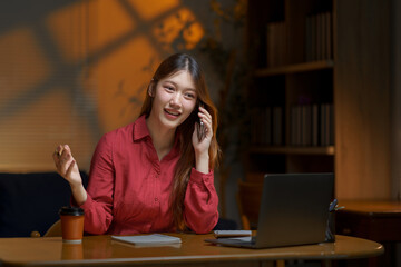 Young woman talking on smartphone and working on laptop at desk in her home office late evening, smiling