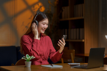 Asian woman relaxing at home, enjoying music or podcast with wireless headphones and smartphone, smiling happily in cozy evening light