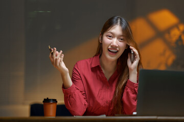 Happy young Asian professional laughing and talking on smartphone at desk, holding a pen while working remotely and planning tasks