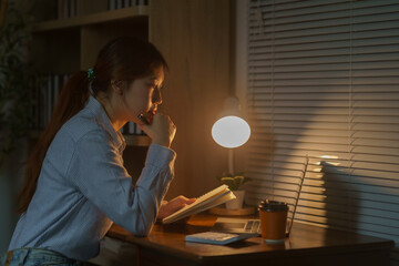 Young woman working or studying, sitting at a desk and using a notebook with a lamp light shining on her face
