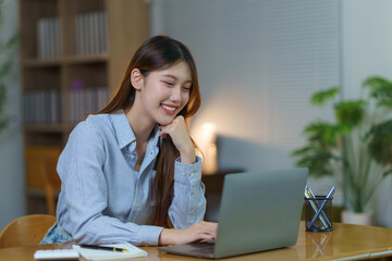Happy Asian woman smiling, using a laptop, and working remotely from her home office, focusing on an online project
