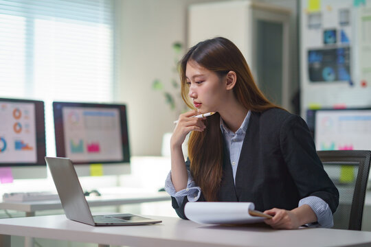 Young Asian businesswoman analyzing data on laptop and holding reports, thinking during a financial review in a modern office