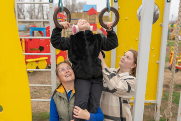 A mother and her friend help a young girl practice exercises on playground rings at an outdoor children's playground, providing support