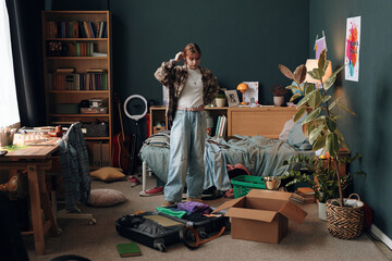 Caucasian teenage girl standing in messy bedroom packing suitcase and cardboard box, organizing clothes and personal items, surrounded by books and plants, appearing thoughtful