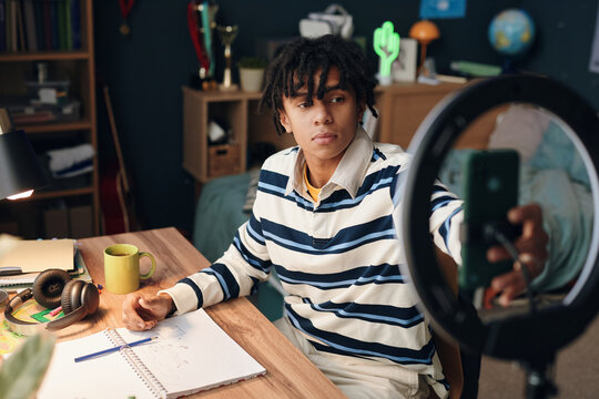 Teenager Black boy sitting at desk recording video on smartphone using ring light, holding phone in one hand while drawing with open sketchbook and pencil in bedroom environment