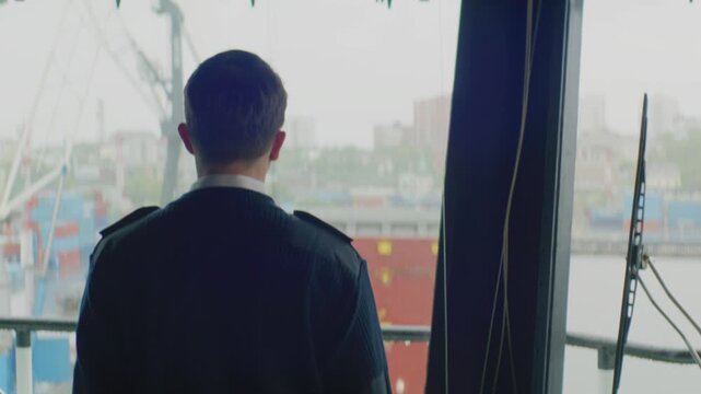 Focused officer observes cranes and ships through foggy glass. Captain stands silently near glass window watching bustling terminal