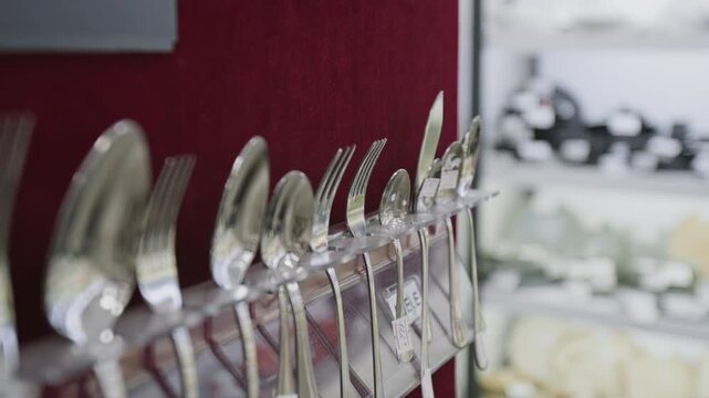 No people close-up of assorted cutlery arranged on red wall display at kitchenware store, steel metal spoons, forks, table knife offered for sale to customers at retail outlet