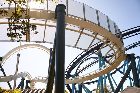 Curving coaster rails pass under a safety net with palms and blue sky. Sunlight and steel shapes create a lively theme park view.