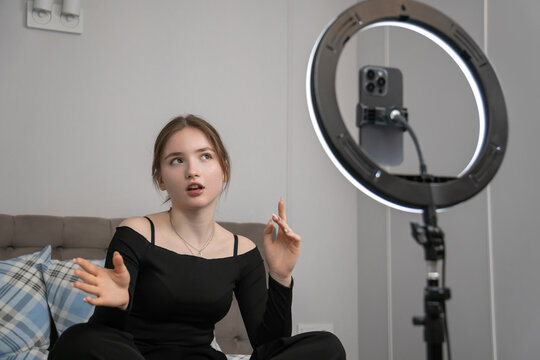 Teenager influencer recording a vibrant vlog while sitting on her bed, using a ring light and smartphone, gesturing animatedly as she engages with her online followers - Powered by Adobe