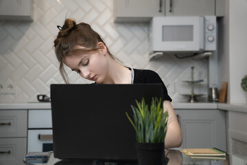 Tired student sitting at desk with laptop after long online learning in home kitchen, feeling tired and overwhelmed with homework and study