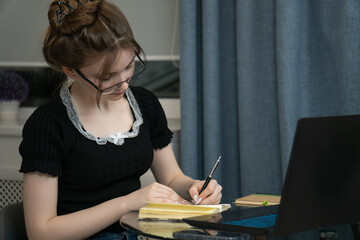 Focused young female student wearing glasses is taking notes in notebook while learning online using laptop, creating comfortable and productive learning space at home