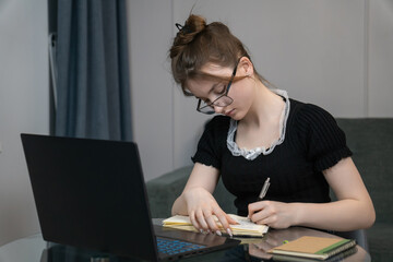 Young girl wearing glasses is taking notes in notebook while learning online using laptop at home, creating comfortable and productive learning environment